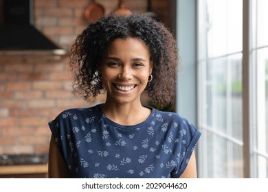 Portrait of happy millennial Black mixed race girl standing by window at home, looking at camera with toothy smile. Beautiful African American young woman, female model in house interior. Head shot - Powered by Shutterstock