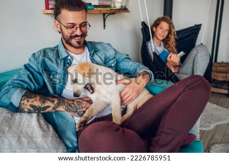 Portrait of happy millennial couple scratching their adopted pet dog and sitting on a sofa at home. Focus on a tattooed man petting their golden retriever in a living room. Copy space.