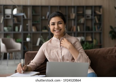Portrait of happy millennial Indian female student use laptop study distant make notes write in notebook. Smiling young mixed race woman work online or take web course on computer at home.