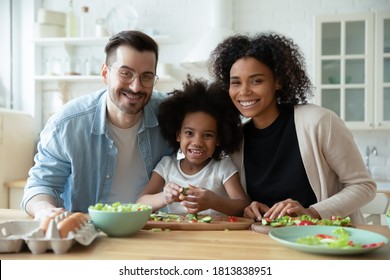 Portrait Of Happy Mixed Race Couple With Small Girl Cooking In Kitchen. Smiling Young Handsome Caucasian Man Enjoying Preparing Food With African Ethnicity Wife And Adopted Little Biracial Daughter.