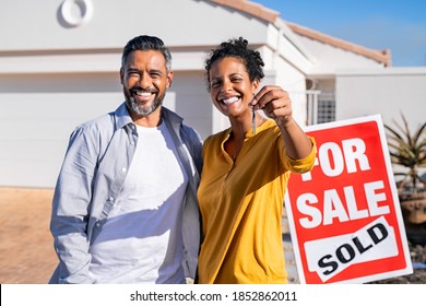 Portrait Of Happy Mixed Race Couple Holding House Keys Standing Near Sold Signboard. Middle Eastern Man Embracing African Woman While Showing House Key Outside Of Their New Home. Moving Home Day.