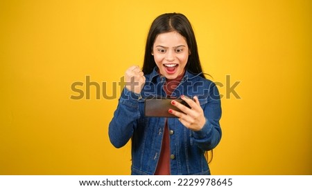 Portrait of happy modern girl using mobile phone, young woman playing video game on smartphone, standing isolated over yellow background