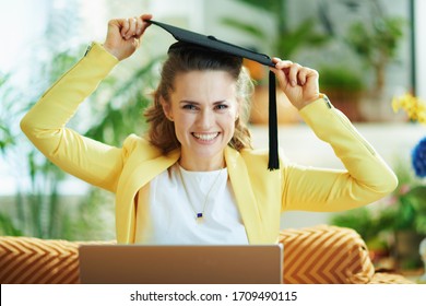 Portrait of happy modern middle age housewife in jeans and yellow jacket with graduation cap and laptop in the modern house in sunny day. - Powered by Shutterstock