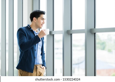 Portrait Of Happy Mood Asian Young Businessman Working Holding A Coffee Cup Wear A Business Suit Of Man In Blue Jacket And Blue Shirt Looking At The Window In The Office Room Background