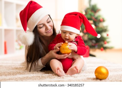 Portrait Of Happy Mother And Adorable Baby Holding Bauble Against Domestic Festive Backdrop With Christmas Tree