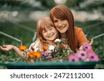 Portrait of happy mother and daughter florists hugging at greenhouse surrounded by flowers.Portrait of cute girl sitting in wheelbarrow with flowers with her mother next to it and smiling at camera.