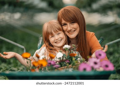 Portrait of happy mother and daughter florists hugging at greenhouse surrounded by flowers.Portrait of cute girl sitting in wheelbarrow with flowers with her mother next to it and smiling at camera. - Powered by Shutterstock