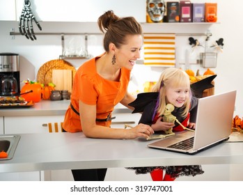 Portrait Of Happy Mother With Daughter In Halloween Bat Costume Having Video Chat On Laptop With Friends In Decorated Kitchen. Traditional Autumn Holiday