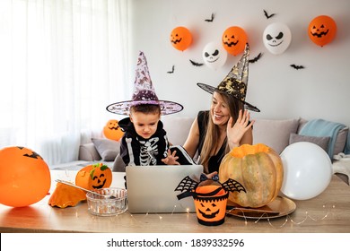 Portrait of happy mother with halloween dressed son having video chat on laptop with friends in decorated livingroom. Traditional autumn holiday. 