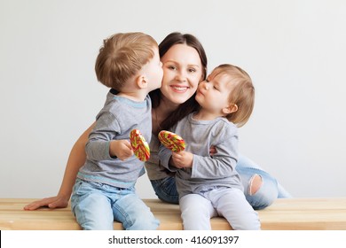 Portrait Of A Happy Mother And Her Two Little Children - Boy And Girl. Happy Family Against A White Background. Little Kids Kissing Mother. Children With Lollipops.