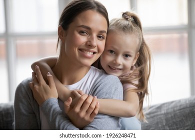 Portrait Of Happy Mother And Little Daughter Kid Hugging On Couch, Looking At Camera, Smiling. Girl And Her Mon Enjoying Being At Home Together, Child Expressing Love And Tender To Her Mum