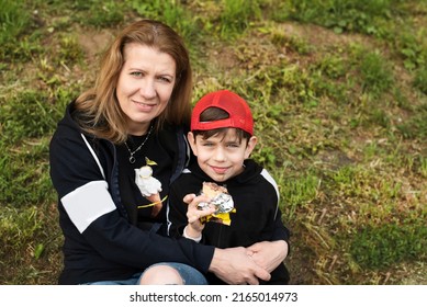 Portrait Of Happy Mother And Little Son, Real Moments Of Life, They Eat Ice Cream And Communicate In The Park Sitting On The Grass, The Boy Has Traces Of Ice Cream On His Face