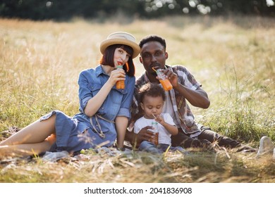 Portrait Of Happy Multi Ethnic Parents Embracing Little Cute Son While Sitting On Wild Field And Drinking Fresh Juice. Family On Picnic. Summer Time Concept.