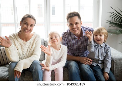 Portrait of happy multi-ethnic family with adopted kids waving hand looking at camera, smiling couple with children sitting on sofa making video call, greeting online by webcam, recording videoblog - Powered by Shutterstock