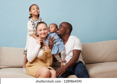 Portrait Of Happy Multiethnic Family Sitting On Couch At Home And Looking At Camera. Black Father, White Mother And Two Daughters. Eldest Leaning On Mother From Behind. Youngest Is In Father's Arms.