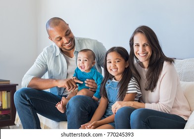 Portrait of happy multiethnic family sitting at home. Smiling couple with kids sitting on couch and looking at camera. Black father and latin woman with daughter sitting on couch and having fun.