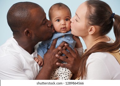 Portrait Of Happy Multiethnic Family: White Mother, Black Father And Their Adorable Little Baby Daughter Against Pale Blue Background. Both Parents Kissing Their Daughter, One Cheek For Each.