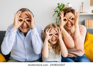 Portrait of happy multi-generational family having fun doing funny faces with fingers sitting together on sofa at home. Cheerful headshot of grandfather, mother and small daugther - Powered by Shutterstock