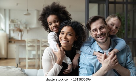 Portrait of happy multiracial couple enjoying sweet family moment with adorable little mixed raced daughters at home. Smiling cute small stepsisters cuddling cheerful parents, looking at camera.