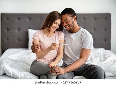 Portrait of happy multiracial couple holding positive pregnancy test, sitting on bed, hugging and enjoying good news. Young loving family rejoycing future parenthood, expecting baby - Powered by Shutterstock