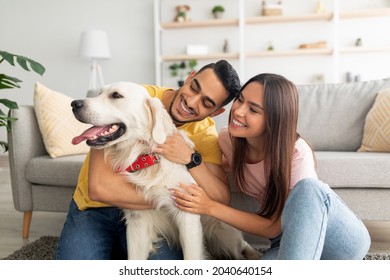Portrait of happy multiracial couple scratching their pet dog, sitting on floor at home. Arab guy and his Caucasian girlfriend hugging their golden retriever in living room