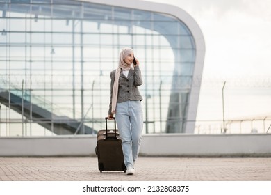 Portrait of happy muslim arab woman wearing a hijab and casual clothes, walking outdoors carrying a suitcase and talking phone, going to travel by airplane at modern airport. - Powered by Shutterstock