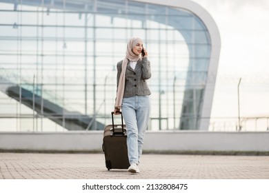 Portrait of happy muslim arab woman wearing a hijab and casual clothes, walking outdoors carrying a suitcase and talking phone, going to travel by airplane at modern airport. - Powered by Shutterstock