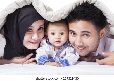 Portrait Of Happy Muslim Family With A Cute Baby Looking And Smiling On The Camera, Shot Under The Blanket On Bed