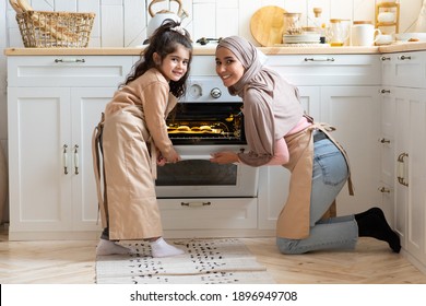 Portrait Of Happy Muslim Family Mother And Daughter Baking Together In Kitchen Interior. Islamic Mom And Her Child Cooking Homemade Pastry, Opening Oven With Freshly Baked Muffins Inside, Free Space