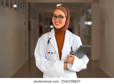 Portrait of happy Muslim female doctor or nurse wearing white coat with stethoscope in hospital corridor holding digital tablet looking at camera and smiling. Professional medical staff headshot. - Powered by Shutterstock