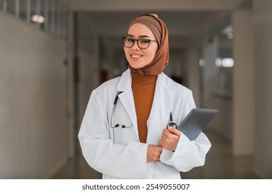 Portrait of happy Muslim female doctor or nurse wearing white coat with stethoscope in hospital corridor holding digital tablet looking at camera and smiling. Professional medical staff headshot. - Powered by Shutterstock