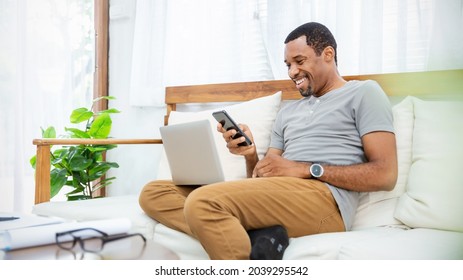 Portrait Of Happy Nerd African Black Man With Casual Cloths Sit On Sofa In Living Room Use Smart Phone Virtual Computer. Cheerful Man Laughing With Phone Online Meeting. Social Media Connected People