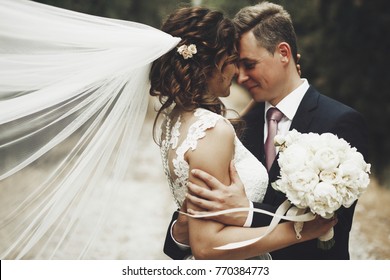 Portrait Of Happy Newly Wife And Husband Hugging Outdoors And Smiling, With Waving Veil And Wedding Bouquet Made Of White Peonies. Sincere Feelings Of Two Young People. Concept Of True Immortal Love.