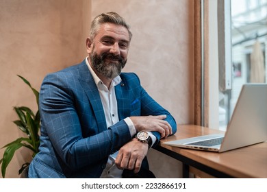 Portrait of a happy old business man sitting at his desk in the office or restaurant cafe near the window and working on a laptop. - Powered by Shutterstock