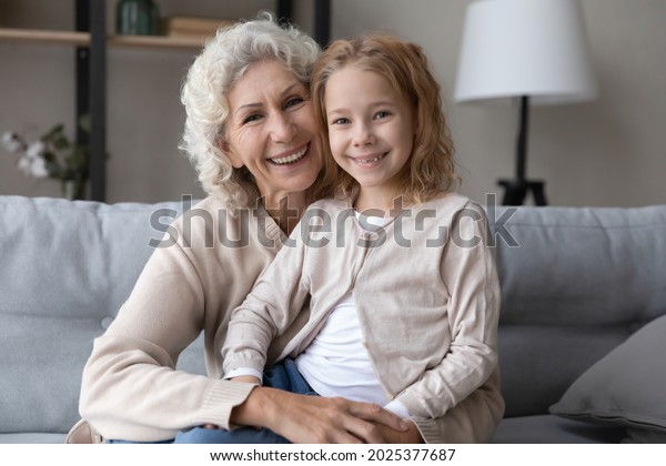Portrait of happy old Caucasian 60s\
grandmother and small teen granddaughter sit rest on sofa at home\
together. Smiling mature grandma and little grandchild cuddle show\
love and care in\
relations.