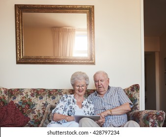 Portrait Of Happy Old Couple Sitting Together At Home On Sofa And Using Digital Tablet. Elderly Man And Woman Using Touchscreen Computer At Old Age Home.