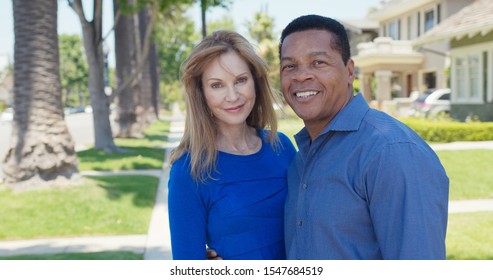 Portrait Of Happy Older African American And Caucasian Couple Smiling On Sidewalk In Suburban Neighborhood. Senior Husband And Wife Homeowners Looking At Camera