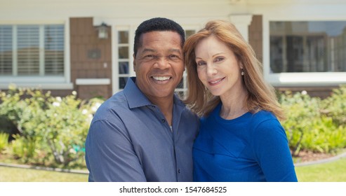 Portrait Of Happy Older African American And Caucasian Couple Smiling In Front Of Their Home. Senior Husband And Wife Homeowners Looking At Camera Outside House