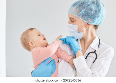 Portrait Of Happy Optimistic Female Doctor Pediatrician Wearing Medical Cap, Gloves And Protective Mask, Standing With Toddler Baby Girl In Hands, Child At Clinic Or Hospital.