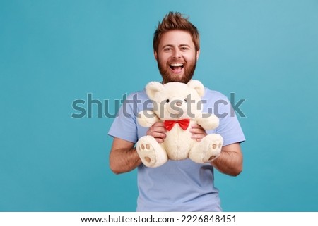 Portrait of happy optimistic handsome bearded man standing looking at camera holding white soft teddy bear, expressing positive emotions. Indoor studio shot isolated on blue background.