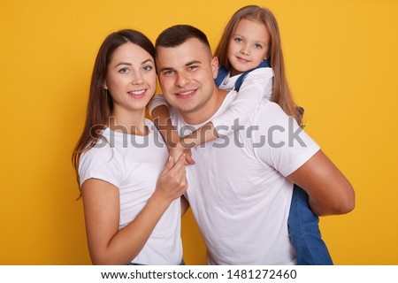 Portrait of happy parents, daddy carries their beautiful daughters on his back. Lovely family of three pose together against yellow background, expressing happuness. Young mother and father with child