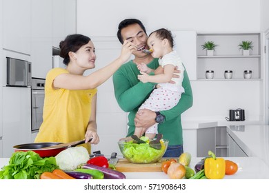 Portrait Of Happy Parents Feeding Their Little Girl While Making Salad In The Kitchen 