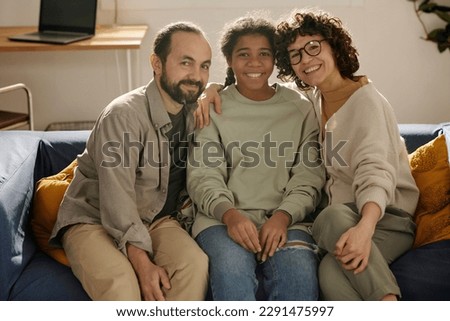 Portrait of happy parents sitting together with their adopted daughter on sofa and smiling at camera