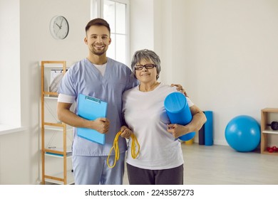 Portrait of happy physiotherapist and senior patient. Young worker with clipboard and old woman with sports equipment standing together in physiotherapy room. Health, exercise, rehabilitation concept - Powered by Shutterstock