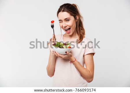 Portrait of a happy playful girl eating fresh salad from a bowl and winking isolated over white background