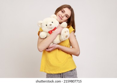 Portrait of happy pleasant looking teenager girl wearing yellow T-shirt hugging romantic present white soft Teddy bear, looks satisfied. Indoor studio shot isolated on gray background.