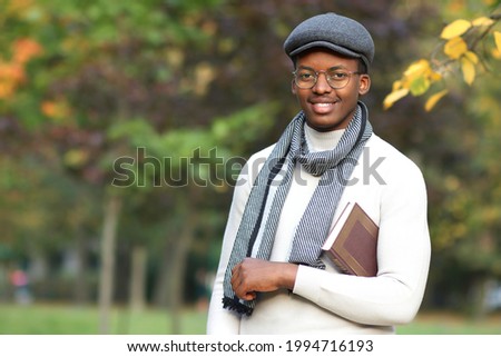Portrait of happy positive black Afro African Afroamerican guy university, college student, handsome ethnic man reader in glasses, scarf, cap with book looking at camera walking in golden autumn park