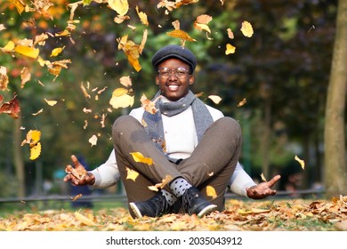 Portrait Of Happy Positive Black Young African Afro American Man Is Sitting Walking, Having Fun In Golden Autumn Park With Falling Colorful Yellow Leaves In Scarf, Hat, Glasses. Beautiful Fall Season