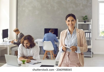 Portrait Of Happy Positive Businesswoman At Work. Cheerful Good Looking Young Woman In Suit Standing In Office, Holding Marker, Looking At Camera And Smiling With Employees Working In Background