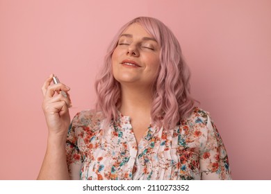 Portrait of happy and positive caucasian female with bottle of perfume applying while standing over bright pink color background. Beautiful girl using perfume